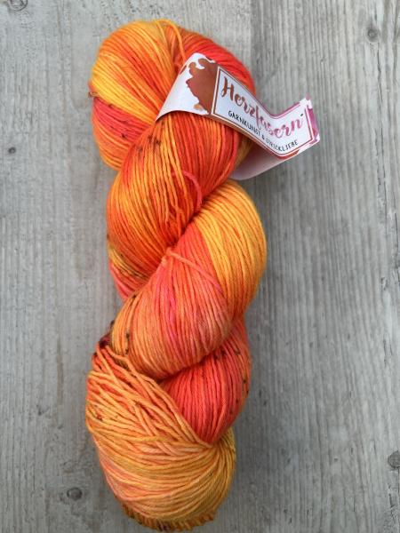 Ein handgefärbter Strang 6-fach Sockenwolle in intensiven Gelb-, Orange- und Pinktönen. Der Strang liegt auf hellem Holz; das Etikett zeigt das Herzfasern-Logo in Pink-Orange.